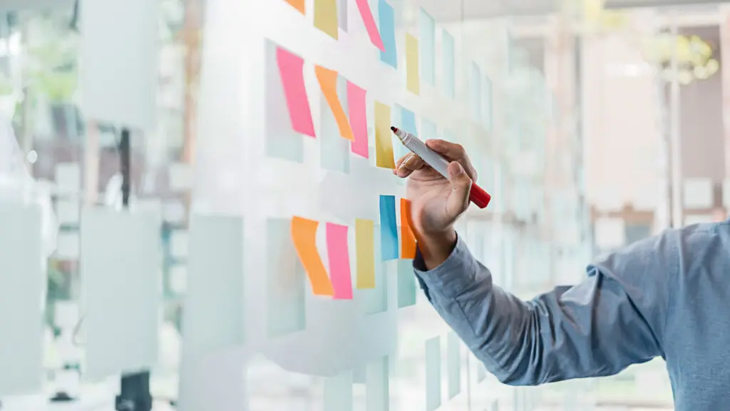 Person writing on colorful sticky notes attached to a glass wall during a brainstorming session
