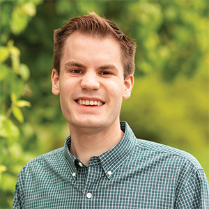 Professional headshot of engineer Andy Zaloudek, wearing a green and white striped button down, standing in front of a leafy background