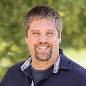Professional headshot of director Ben Hayward wearing a blue button down over a gray t-shirt, standing in front of a leafy background