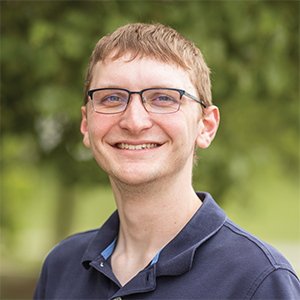 Professional headshot of engineer, Ben Wittrup, wearing a navy blue polo, standing in front of a leafy background