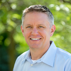 Professional headshot of founding and Managing Partner Casey Alford wearing a blue polo shirt against a leafy green background.