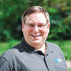 Professional headshot of engineer Chris Danks, wearing a gray polo with the Treetown Tech logo on the left side, standing in front of a leafy background