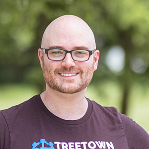 Professional headshot of Chris Eckmeter, wearing a burgandy Treetown Tech shirt, standing in front of a leafy background
