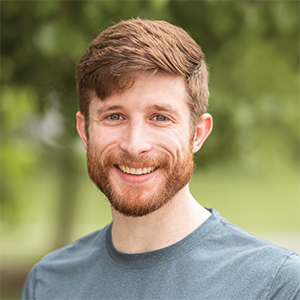 Professional headshot of engineer Cody Mulhollen wearing a grey t shirt, standing in front of a leafy background