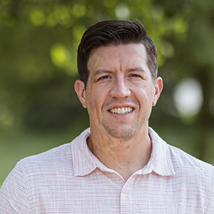 Professional headshot of Mechanical Engineering expert, Chad Neumann, wearing a white button down, standing in front of a leafy background