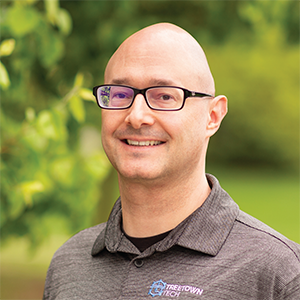 Professional headshot of engineer, Chris Silkowski, wearing a heather gray Treetown Tech polo over a black t-shirt, standing in front of a leafy background