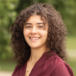 Professional headshot of engineer Elena DeLaCruz wearing a burgandy blouse, standing in front of a leafy background