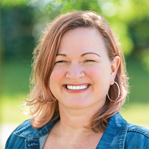 Professional headshot of operations director, Esther Franck, wearing a blue jean button up, standing in front of a leafy background