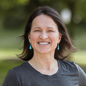 Profesisonal headshot of Elena Haviland wearing a gray t-shirt, standing in front of a leafy background