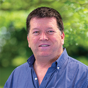 Professional headshot of engineer Greg Robertson wearing a blue button down, standing in front of a leafy background