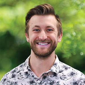Professional headshot of engineer Guerin Rowland,wearing a black and white patterned button down, standing in front of a leafy background