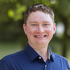 Professional headshot of James Coller wearing a blue button down standing in front of a leafy background