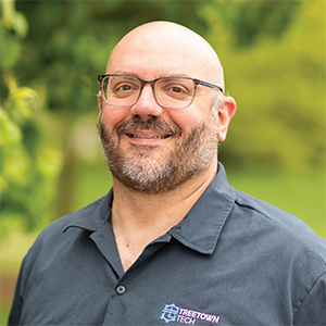 Professional headshot of engineer Joe DiGiglio wearing a blue Treetown Tech dickies cover shirt, standing in front of a leafy background.