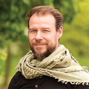 Professional headshot of director Justin Tesmer, wearing an olive green afghan around his neck, over a black t-shirt, standing in front of a leafy background