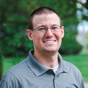 Professional headshot of engineer Kevin Bancroft, wearing a gray Treetown Tech polo, with a white t-shirt underneath, standing in front of a leafy background