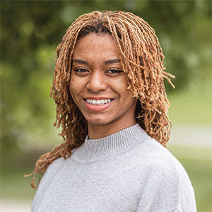Professional headshot of engineer Kamayah Carr-Bunkley wearing a gray turtleneck sweater, standing in front of a leafy background