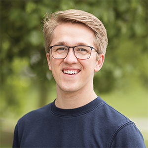 Professional headshot of engineer Kyle Coon wearing a navy blue crew neck, standing in front of a leafy background