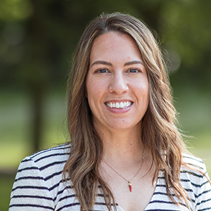 Professional headshot of UI/UX designer Kristen Greco, wearing a striped blouse, standing in front of a leafy background