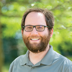 Professional headshot of founding partner, Kris Schilling, wearing a greenish grey button down, standing in front of a leafy background