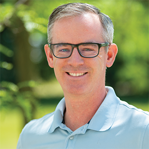 Professional headshot of director Larry Mercier, wearing a blue polo, standining in front of a leafy background