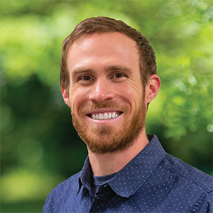 Professional headshot of project manager, Luke Paul, wearing a dark blue button down shirt, standing in front of a leafy background