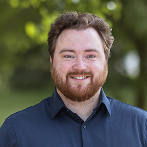 Professional headshot of Project Manager Matt Giles wearing a blue button down, standing in front of a leafy background