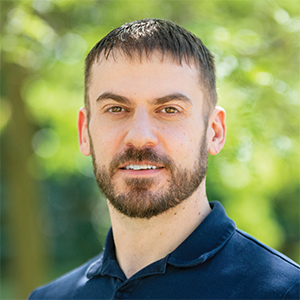 Professional headshot of founding partner, Mike Gorski, wearing a dark blue polo, standing in front of a leafy background