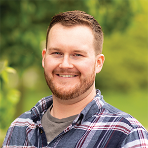 Professional headshot of fabricator Michael Mosher wearting a blue plaid button down over a gray t-shirt, standing in front of a leafy background