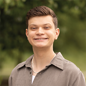 Professional headshot of engineer Nick Antony, wearing a brown button down over a white shirt, standing in front of a leafy background
