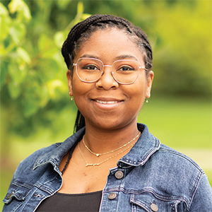 Professional headshot of Noelle Douglas, wearing a dark blue jean jacket over a black camisol, standing in front of a leafy green background