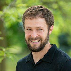 Professional headshot of founder, Nate Ernst, wearing a black polo, standing in front of a leafy background