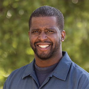 Professional headshot of fabricator Pierre Coward wearing an indigo dickies cover shirt over a dark blue t-shirt, stranding in front of a leafy background