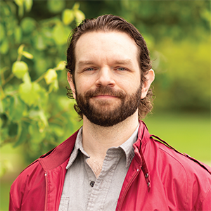 Professional headshot of Ryan Brzys wearing a red bomber jacket over a tan button down, standing in front of a leafy background