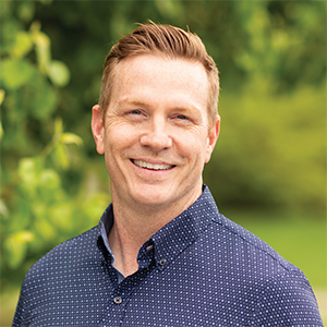 Professional headshot of engineer Scott Campbell wearing an indigo button down, standing in front of a leafy background